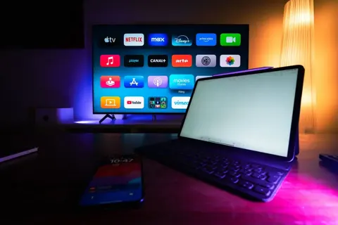 Televisão com aplicativos e notebook aberto em ambiente com iluminação colorida.