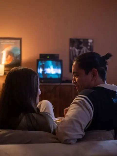 Casal no sofá diante de televisão em sala com iluminação noturna.